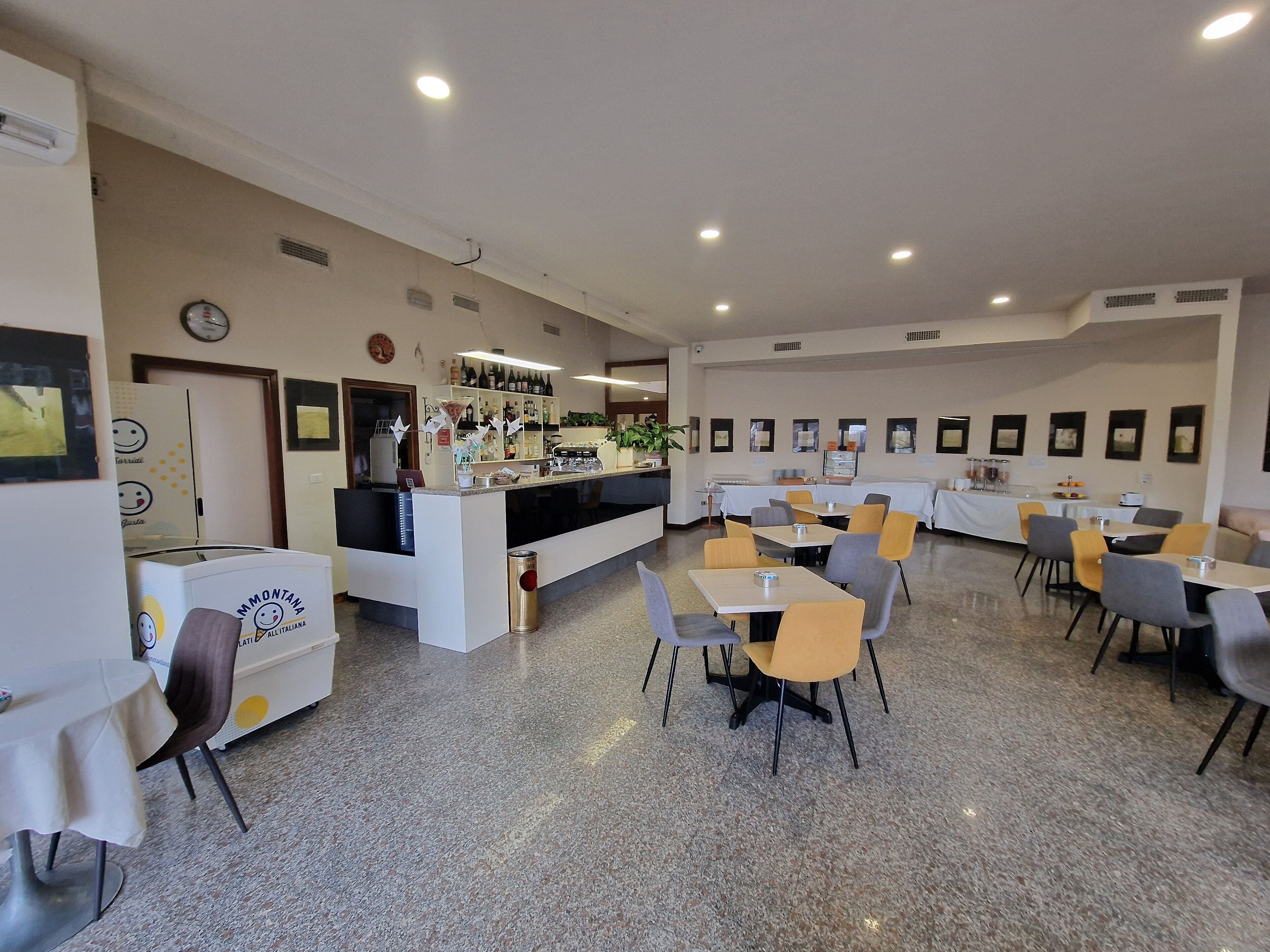 Un vaste espace dédié à la restauration, avec de nombreuses tables et chaises, un comptoir d’accueil et un bar où sont exposées des bouteilles et des verres.