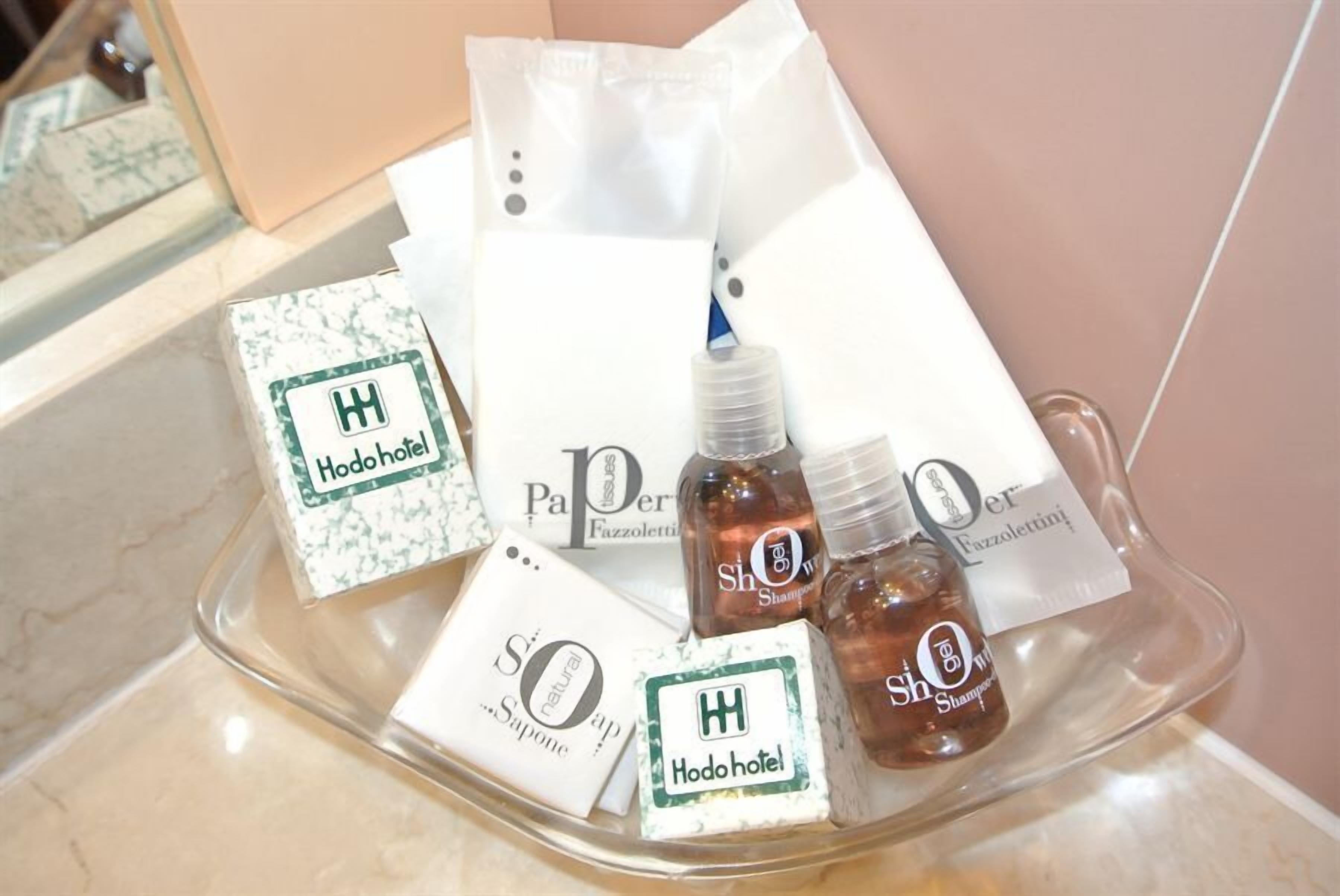 Uma bandeja com amenities de higiene pessoal, incluindo uma garrafa de shampoo, um sabonete em barra e um cartão com o logotipo do hotel Hodo.