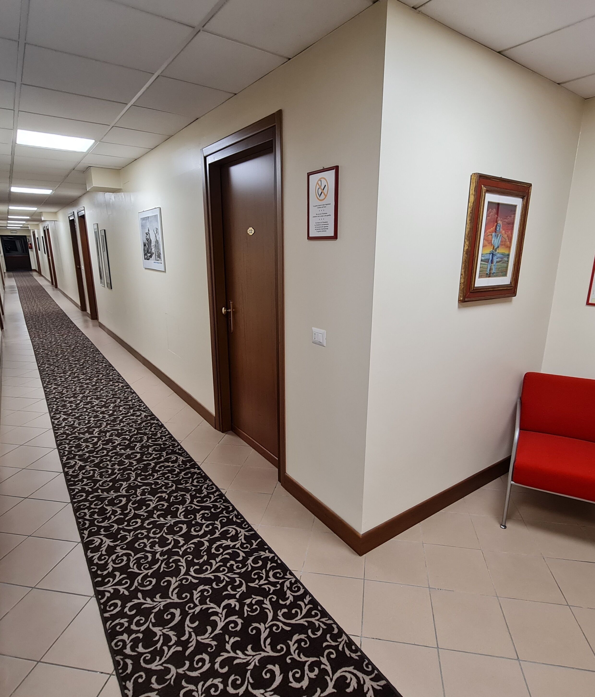 A hotel corridor with a patterned carpet, a red bench, and framed artwork on the walls.