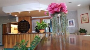 A reception area with a wooden counter, a large decorative wall piece, potted plants, and framed artwork on the wall. - Hotel Passacor (Borgocarbonara)