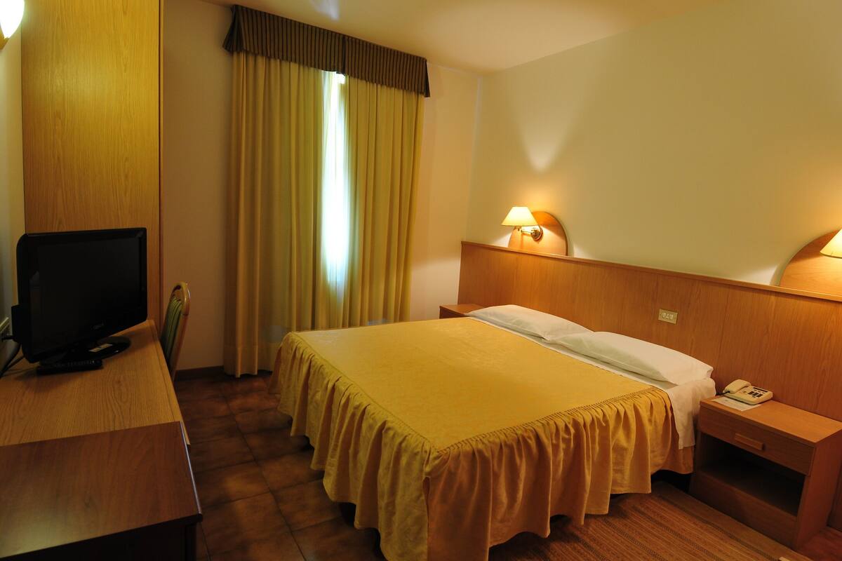 A hotel room with a bed, a desk with a computer, a nightstand with a lamp, and a window with curtains.