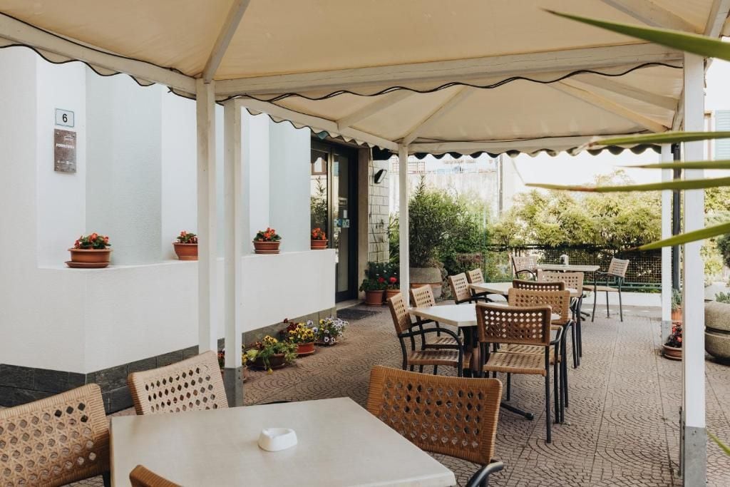 Un patio al aire libre con mesas y sillas, rodeado de plantas en macetas y una pared blanca con un letrero con número.