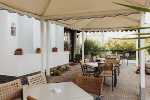 An outdoor patio with tables and chairs, surrounded by potted plants and a white wall with a number sign.
