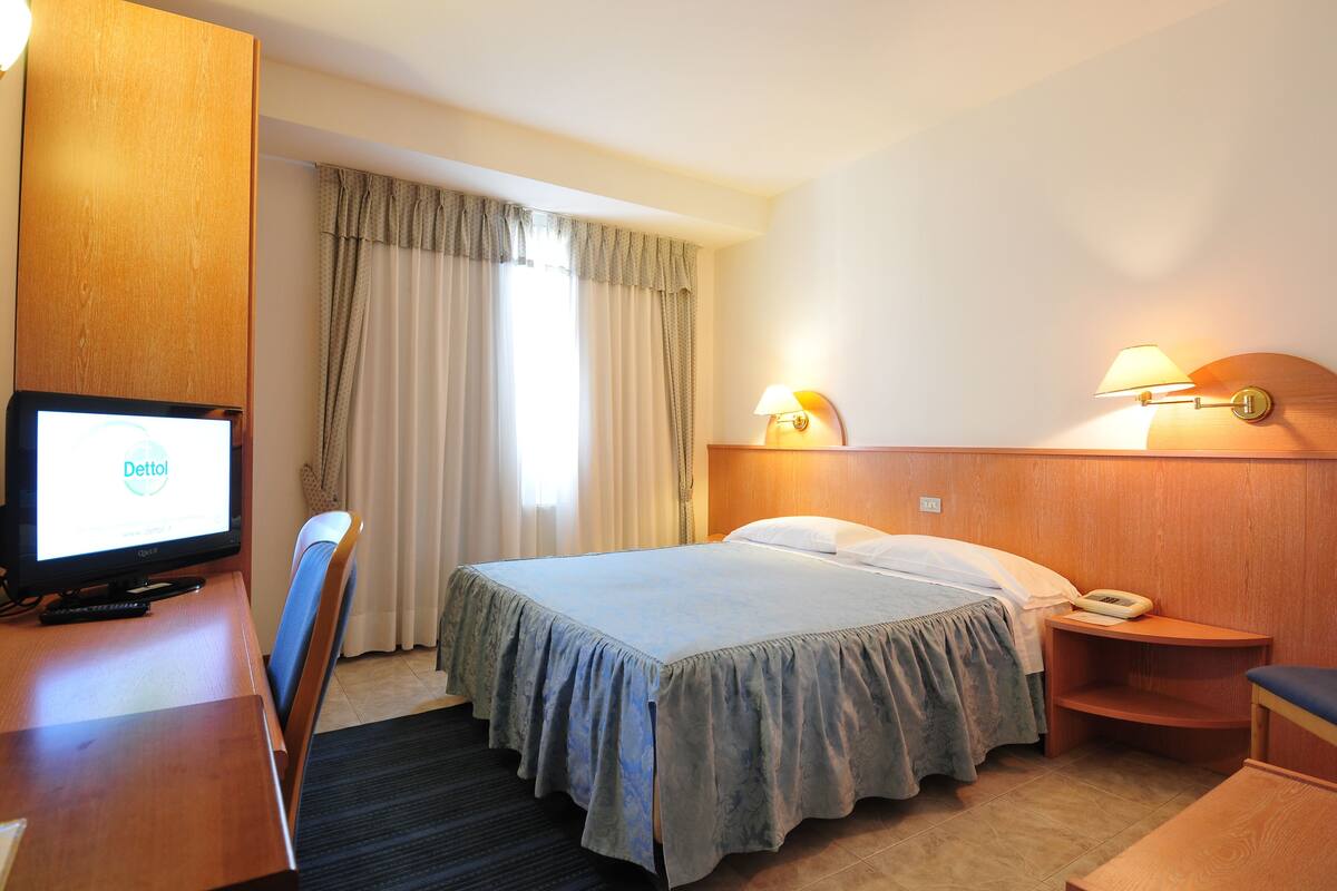 A hotel room with a bed, a desk with a computer, a chair, a bedside table with a lamp, and a window with curtains.