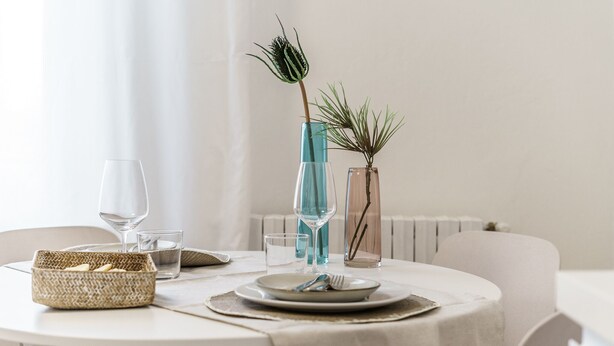 Ein runder Esstisch mit weißer Tischdecke, ein gewebter Korb, ein Glas Wasser, ein Teller mit Besteck und zwei Glasvasen mit Pflanzen.