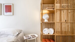 Ein ordentlich eingerichtetes Hotelzimmer mit einem Bett, einem kleinen Tisch mit einer Zeitschrift, einem Holzregal mit Handtüchern und einem gerahmten Bild an der Wand.