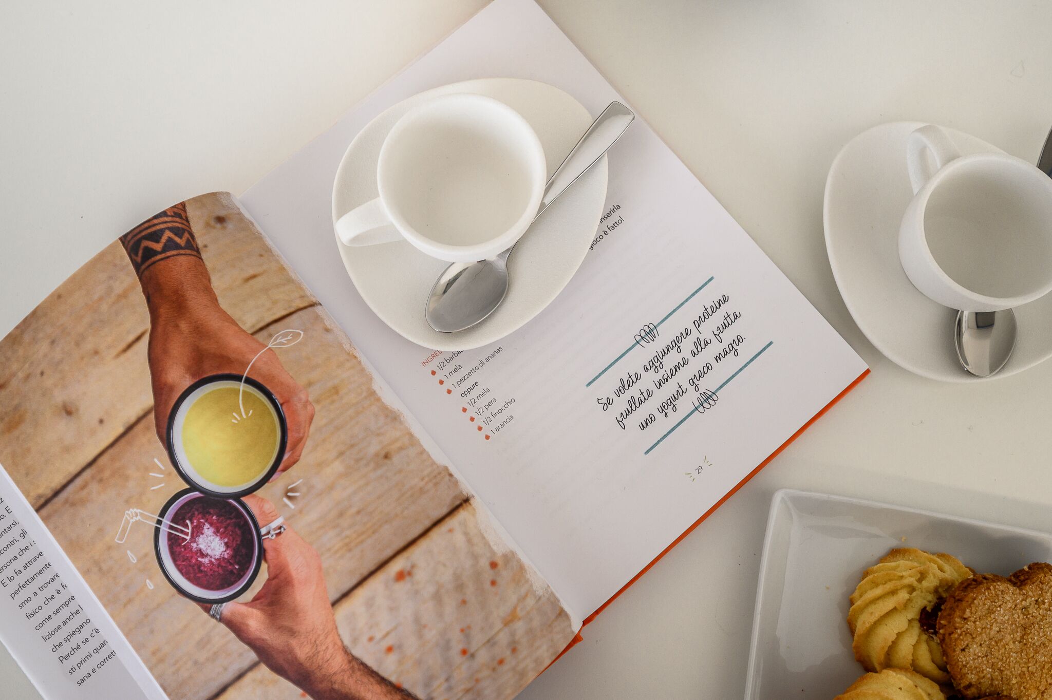A book open on a table with a cup and saucer, a spoon, and a plate with pastries.