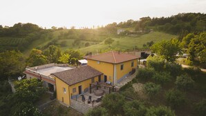 Un bâtiment jaune, surmonté d’un toit en tuiles, entouré de verdure et de collines.