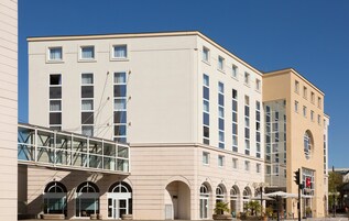 Un hotel moderno con fachada color beige, múltiples ventanales y un puente de cristal que lo conecta a otro edificio.