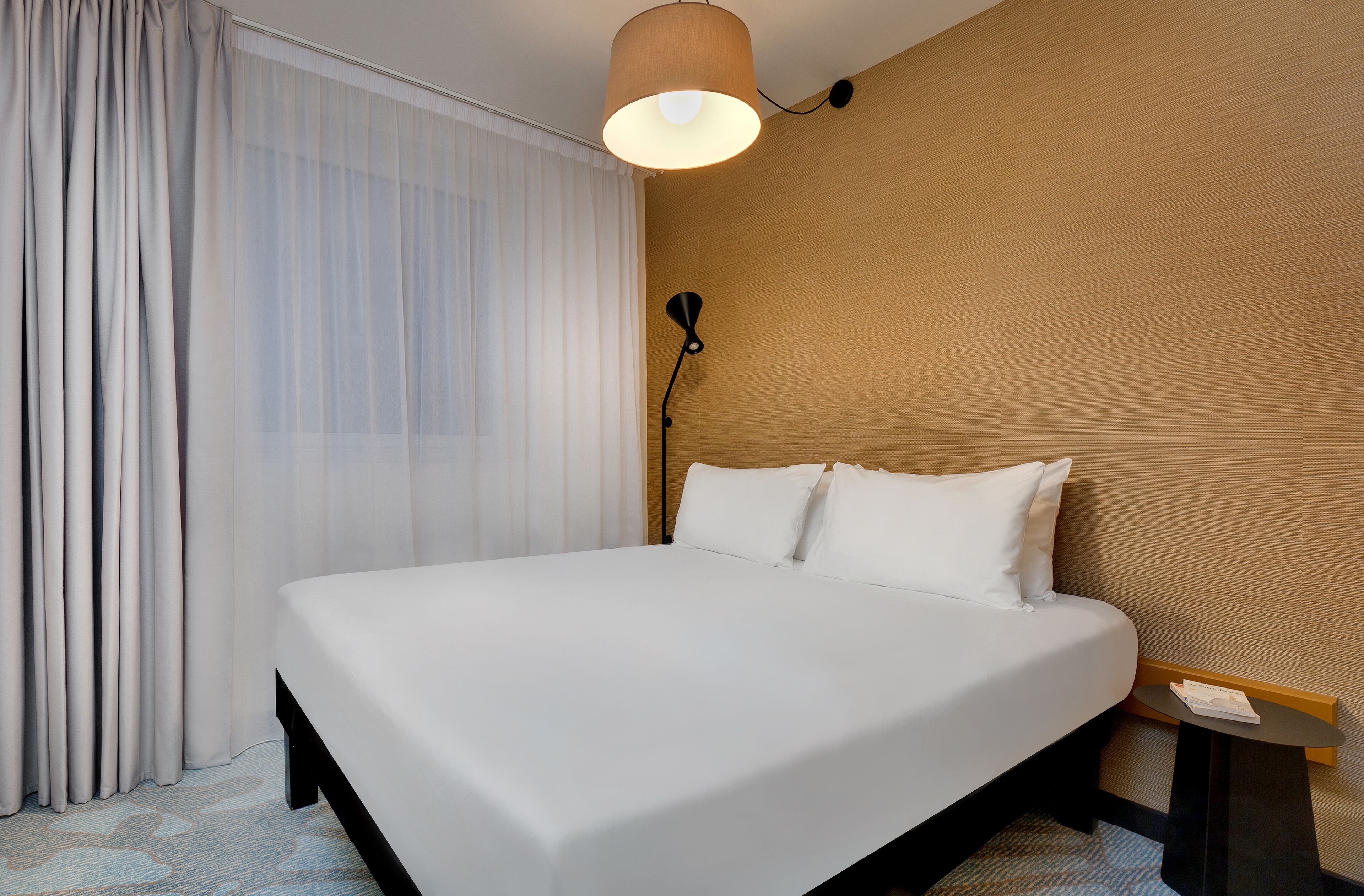 A hotel room with a large bed, a bedside table, a lamp, and sheer curtains.