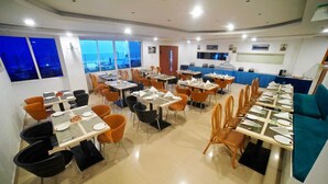 A dining area with multiple tables and chairs, large windows with a view, and a counter with a blue tablecloth. - Bay View Hotel (Visakhapatnam)