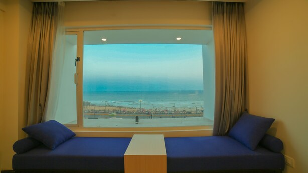 A room with a large window overlooking a beach, a blue sofa with cushions, and a wooden table in the center. - Bay View Hotel (Visakhapatnam)