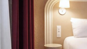 Una habitación de hotel con cama, lámpara de noche, mesita de noche y ventana con cortinas.