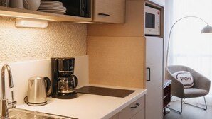 Cocina moderna con un lavamanos de acero inoxidable, una cafetera y un microondas. En la habitación contigua hay una silla con un cojín que dice "VICHY".