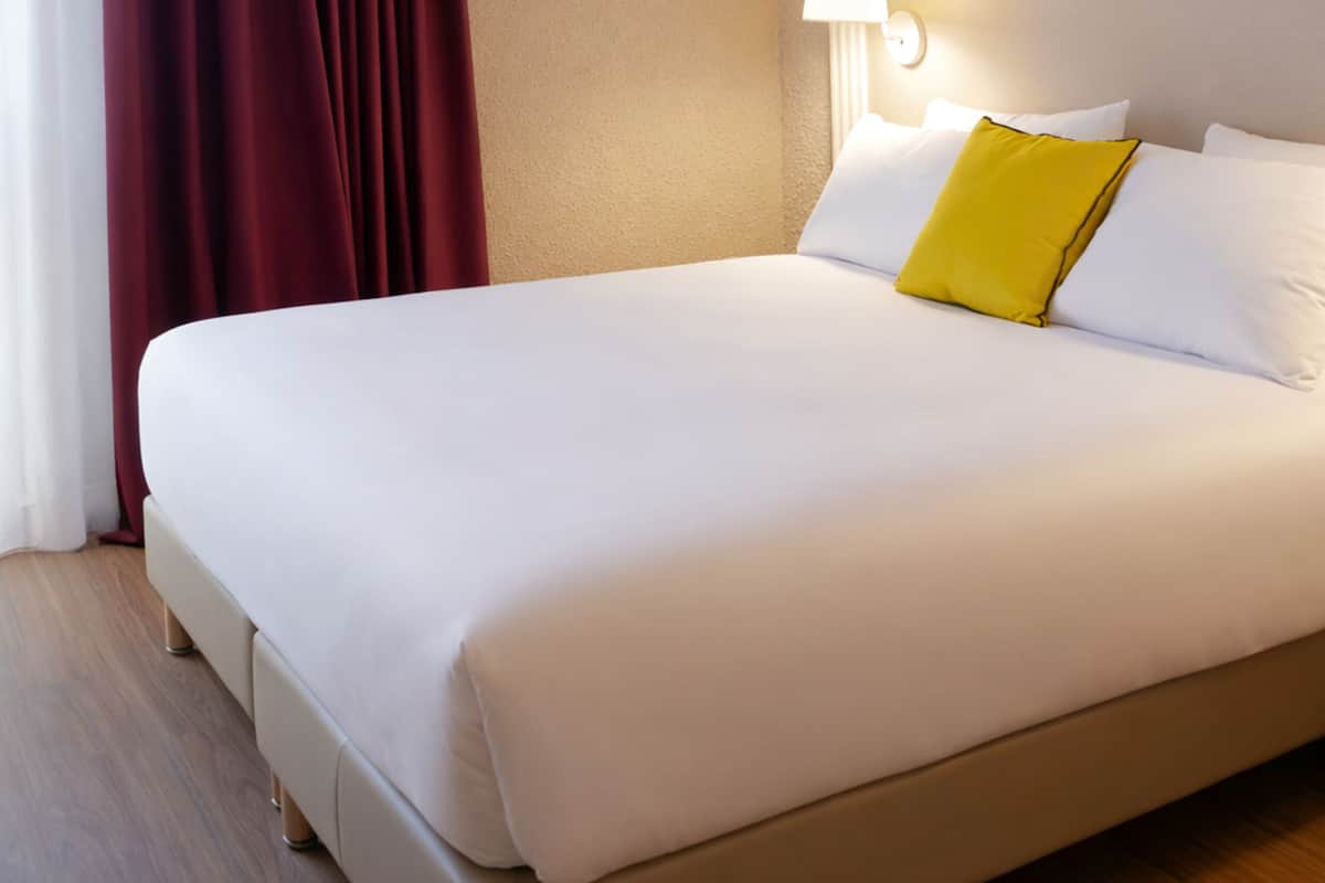Un lit bien fait, avec des oreillers blancs et jaunes, une tête de lit beige et un rideau rouge.