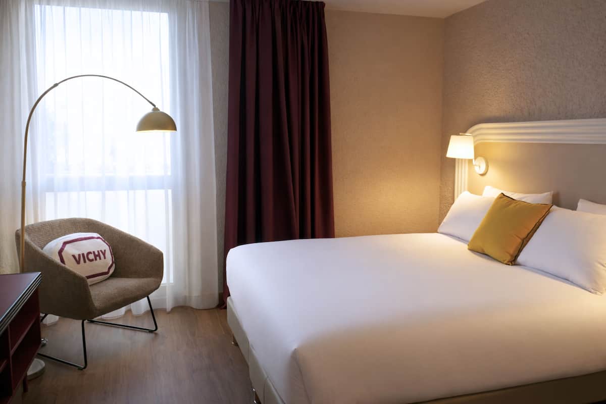 Une chambre d’hôtel avec un grand lit, une chaise avec un coussin portant l’inscription « VICHY », un bureau et une lampe.