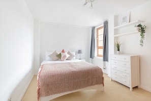 A bedroom with a bed, a dresser, a window with curtains, and a shelf with a plant. - Serenity Apartment Whitechapel (London)