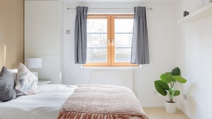 A bedroom with a bed, a nightstand, a window with curtains, a radiator, and a potted plant. - Serenity Apartment Whitechapel (London)