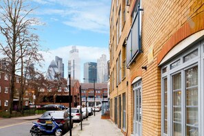 A narrow urban street with parked cars, a brick building with large windows, and a city skyline in the background.