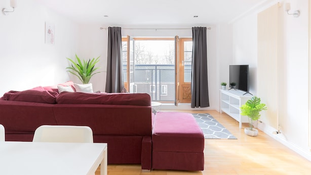 A modern living room with a red sofa, a white table, a TV, and a balcony with curtains. - Serenity Apartment Whitechapel (London)