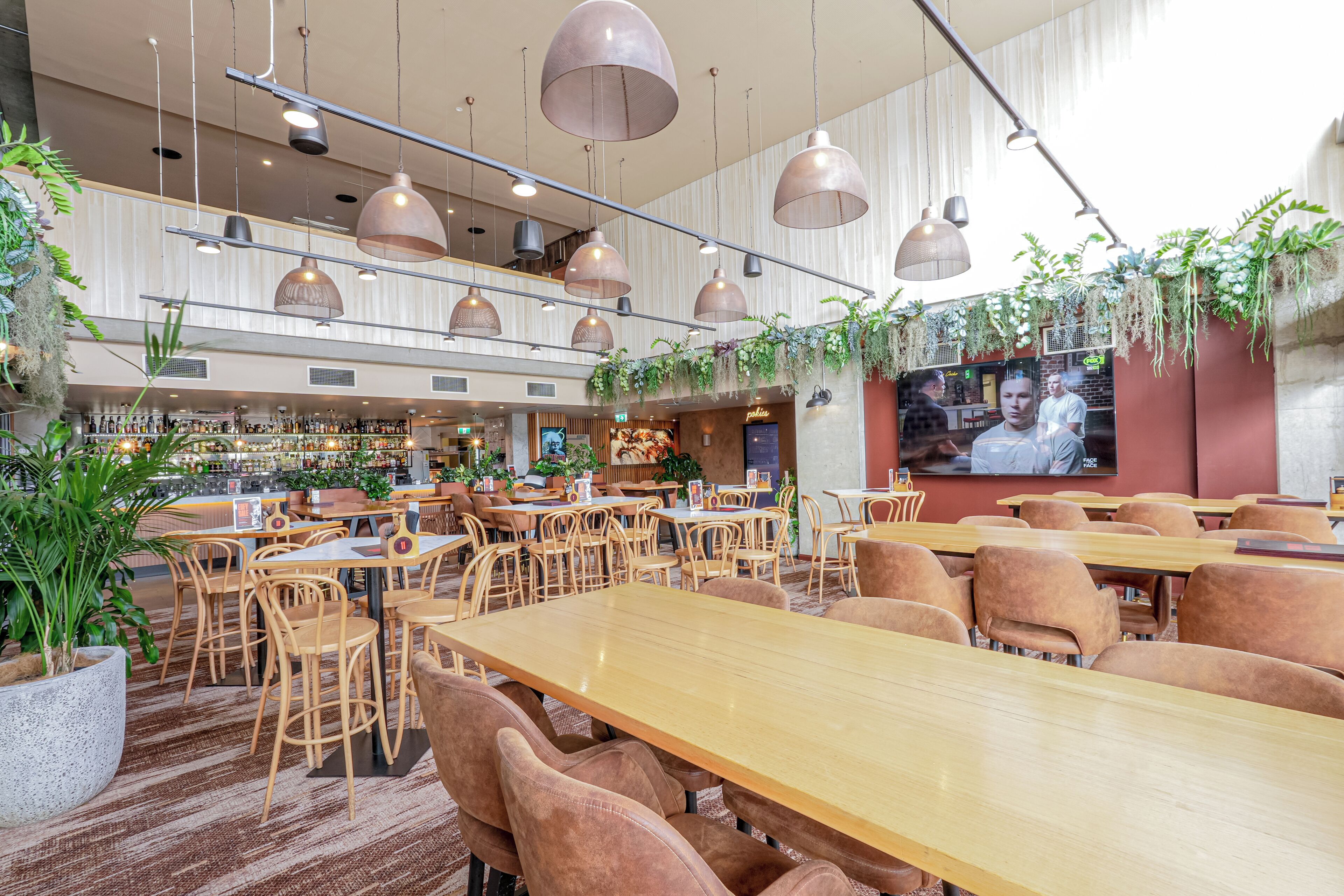 Un restaurante con varias mesas y sillas, una zona de bar con estantes llenos de botellas y plantas colgantes.