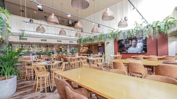 Un restaurante con varias mesas y sillas, una zona de bar con estantes llenos de botellas y plantas colgantes.