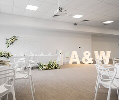 Una habitación amplia con una larga mesa dispuesta para un evento formal, sillas blancas y letras iluminadas de gran tamaño que deletrean “A&W” en la pared.
