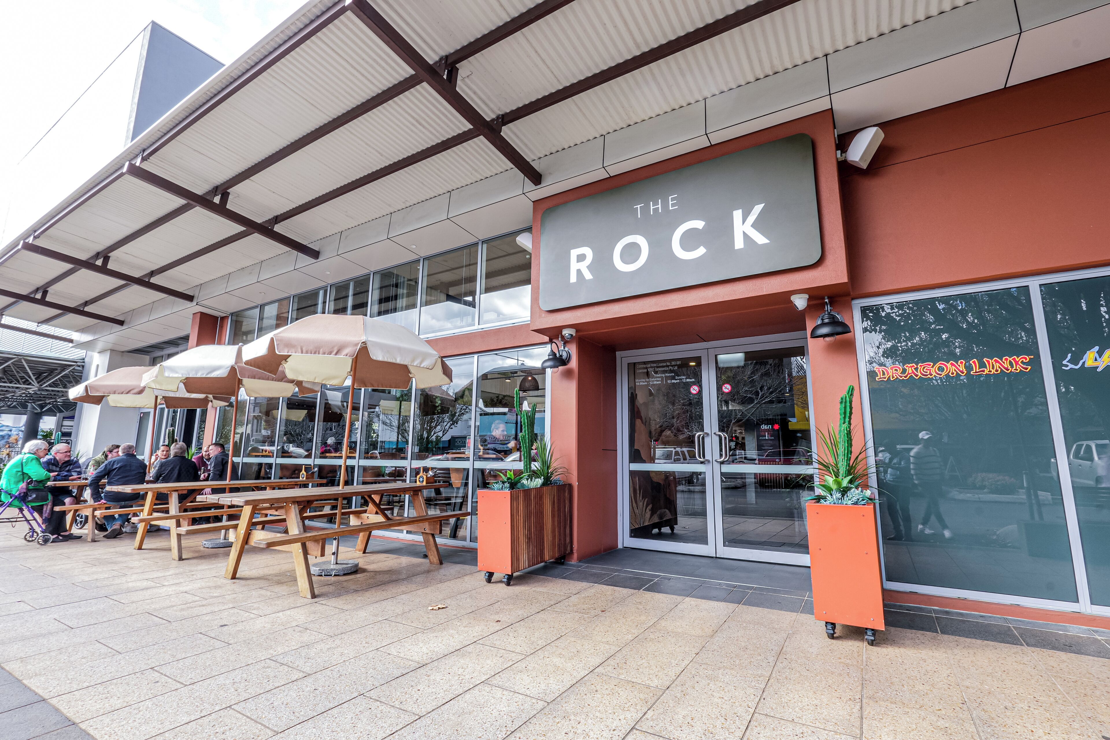 An outdoor seating area with wooden tables and umbrellas, a storefront with a sign reading 'The Rock', and a glass door entrance.