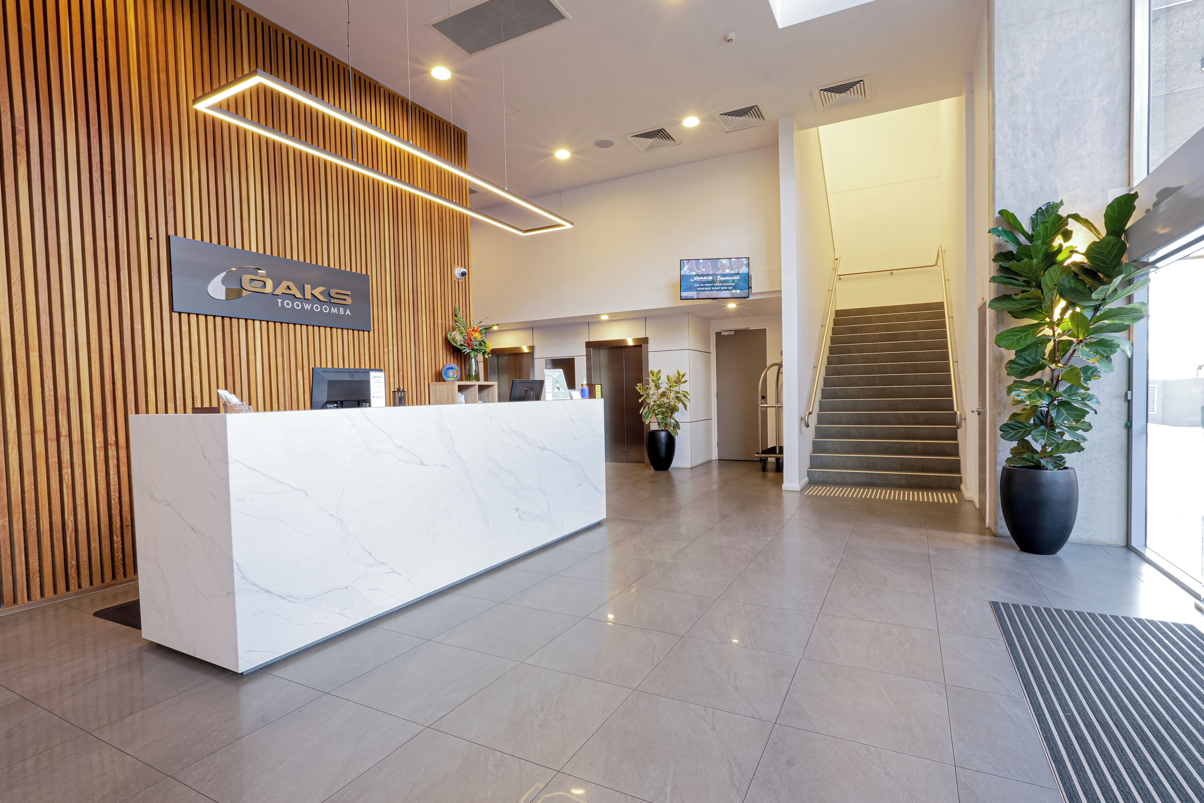 A hotel reception area with a modern design, featuring a reception desk, a wooden wall, a large potted plant, and a staircase.