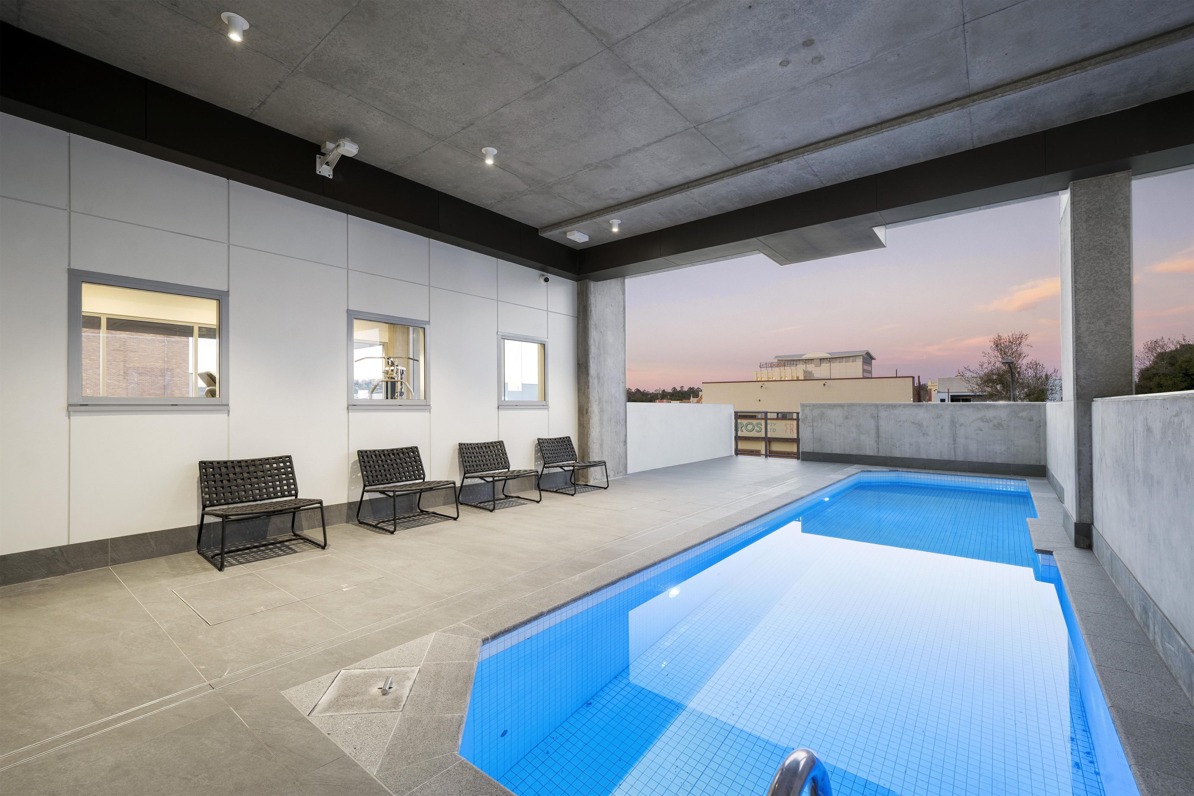 An indoor swimming pool with a tiled floor, benches along the wall, and a view of the cityscape at dusk.