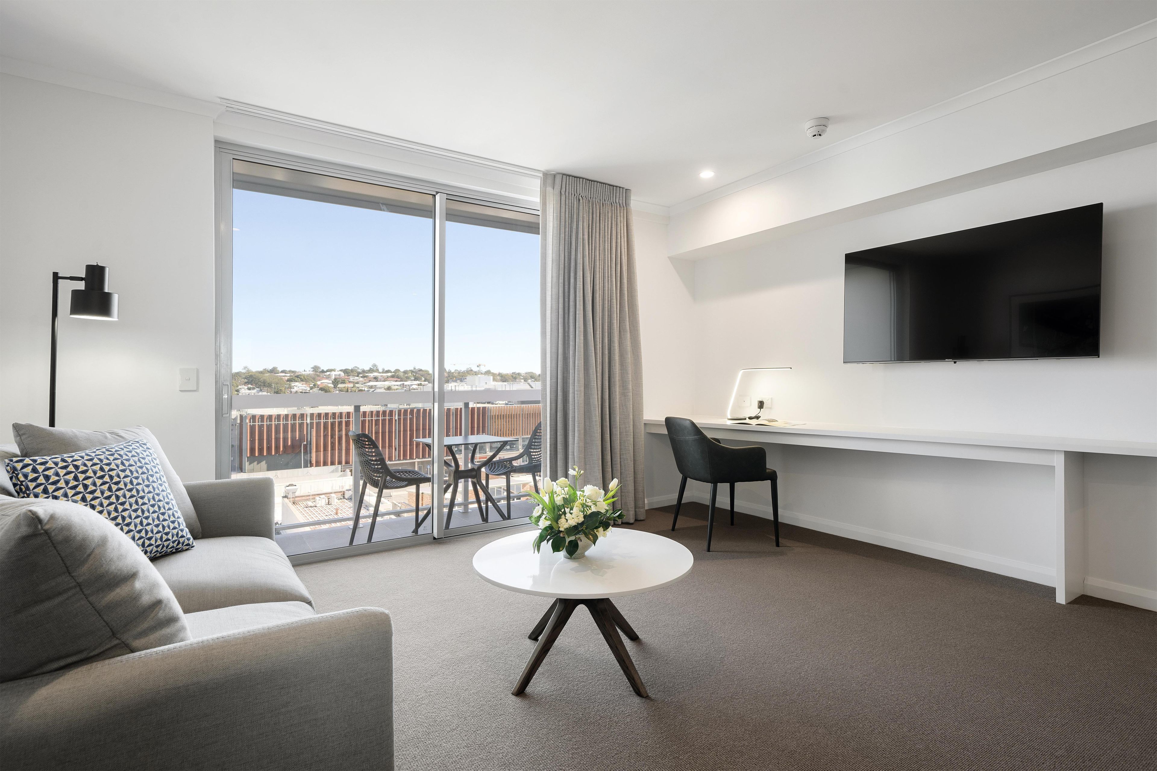 A modern hotel room with a sofa, a desk, a TV, and a balcony with outdoor seating.