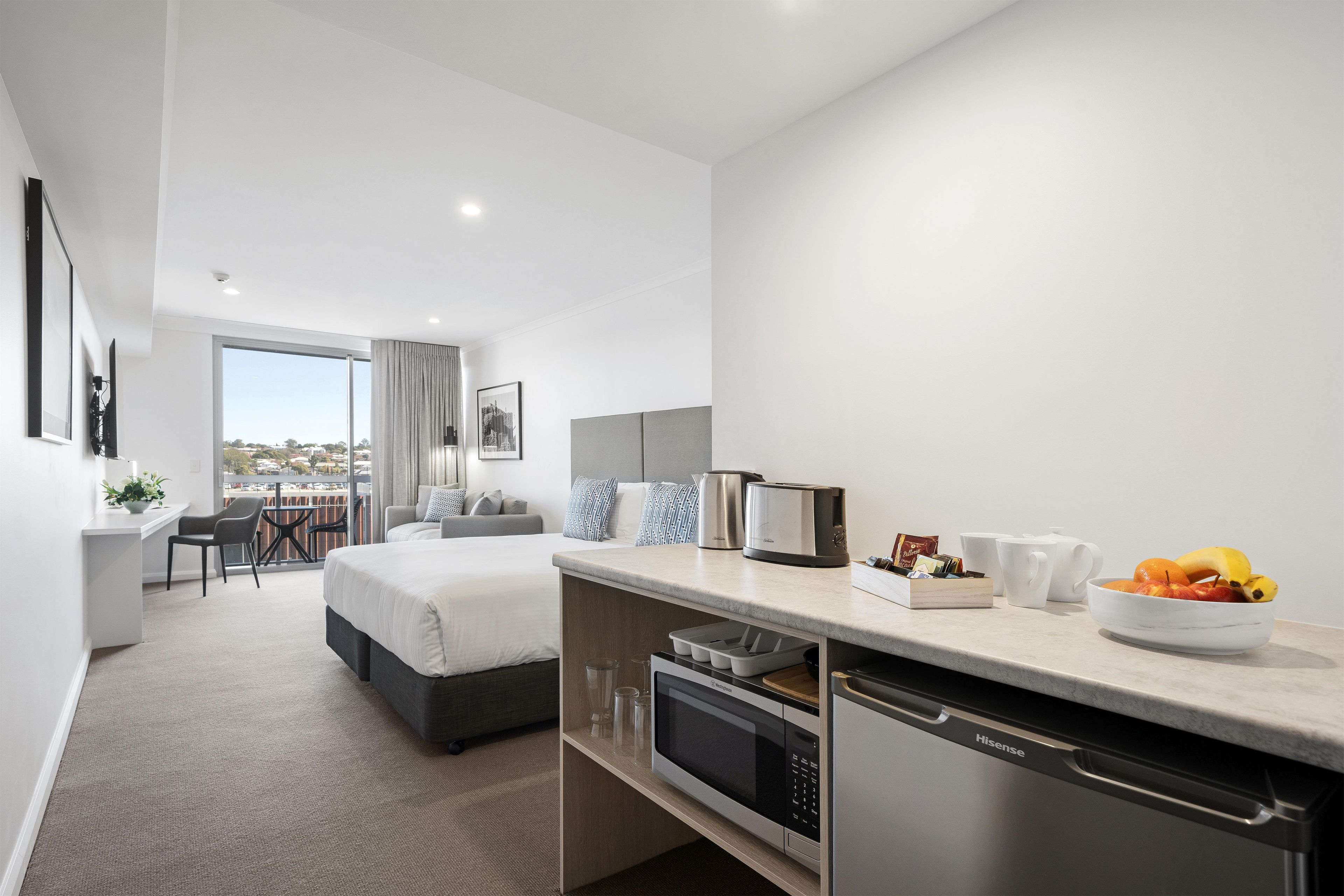 A modern hotel room with a bed, a seating area, a kitchenette, and a view of the city.