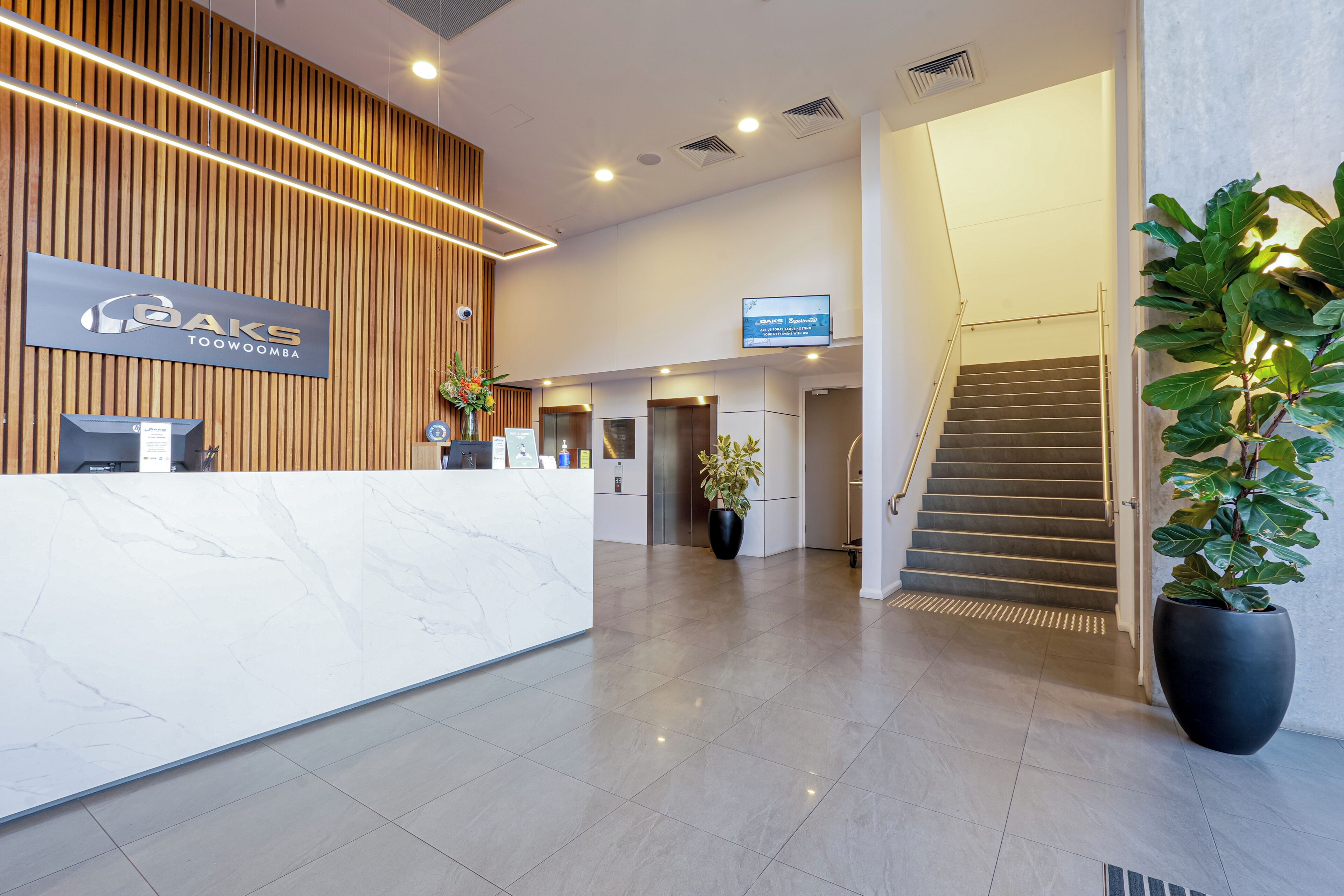 A hotel reception area with a modern design, including a reception desk, a digital screen, and a staircase.