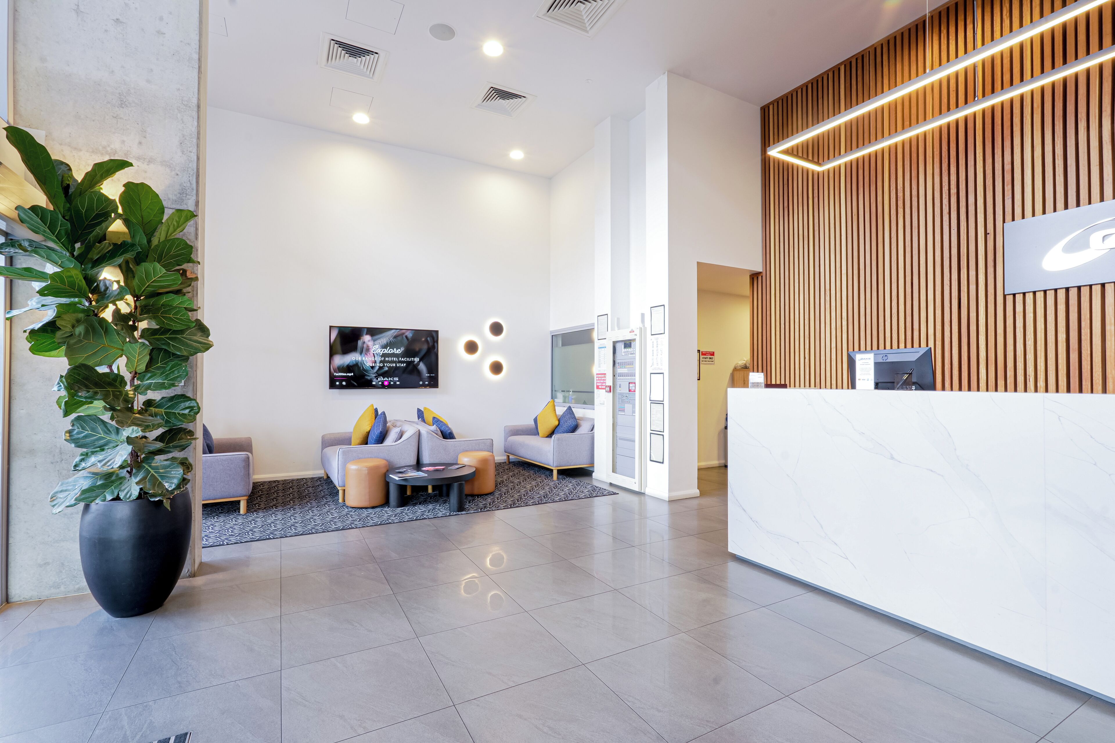 A modern hotel reception area with a white reception desk, a large potted plant, and a seating area with sofas and a coffee table.
