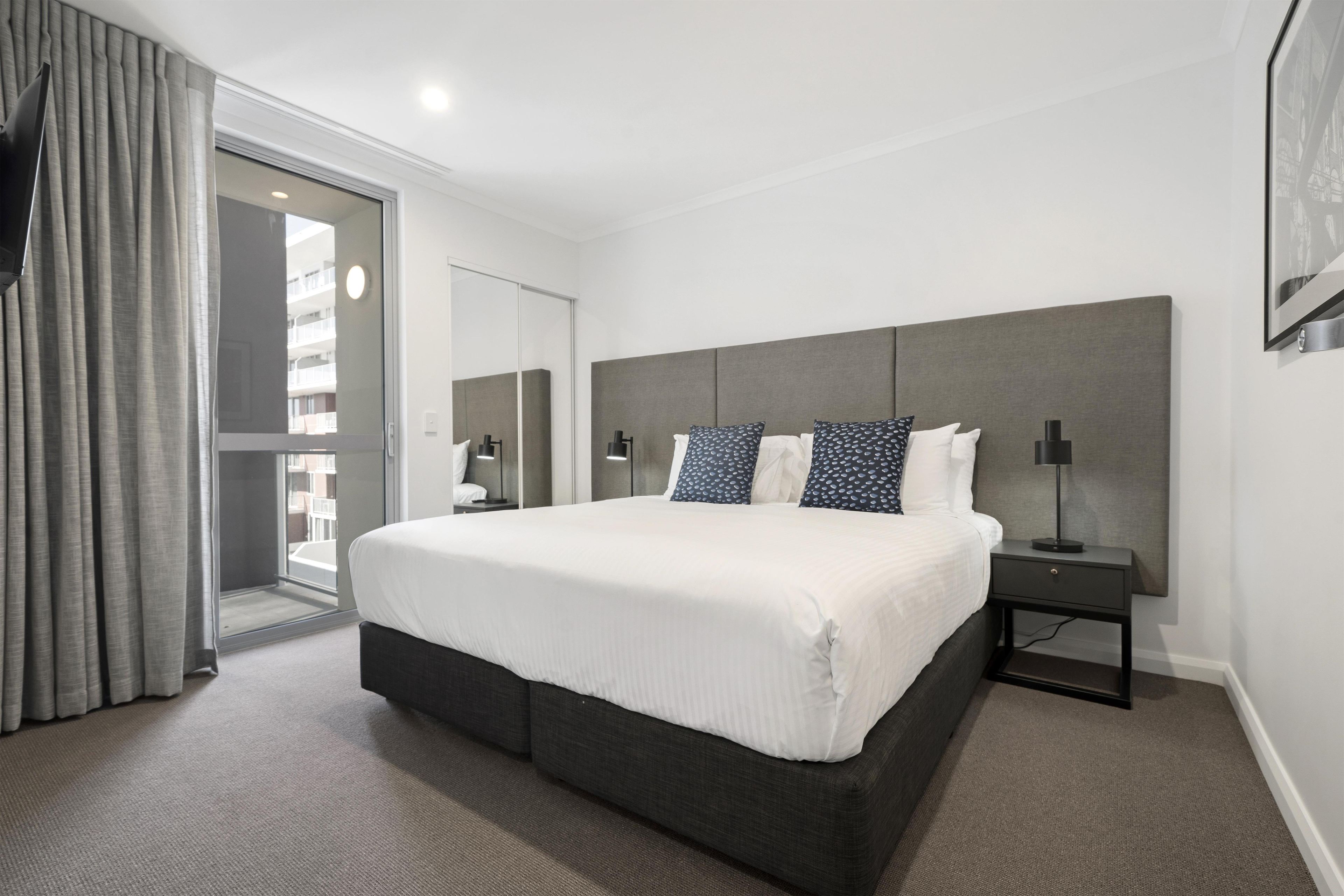 A hotel room with a large bed, a bedside table, a nightstand, and a view of the outside through a sliding door.
