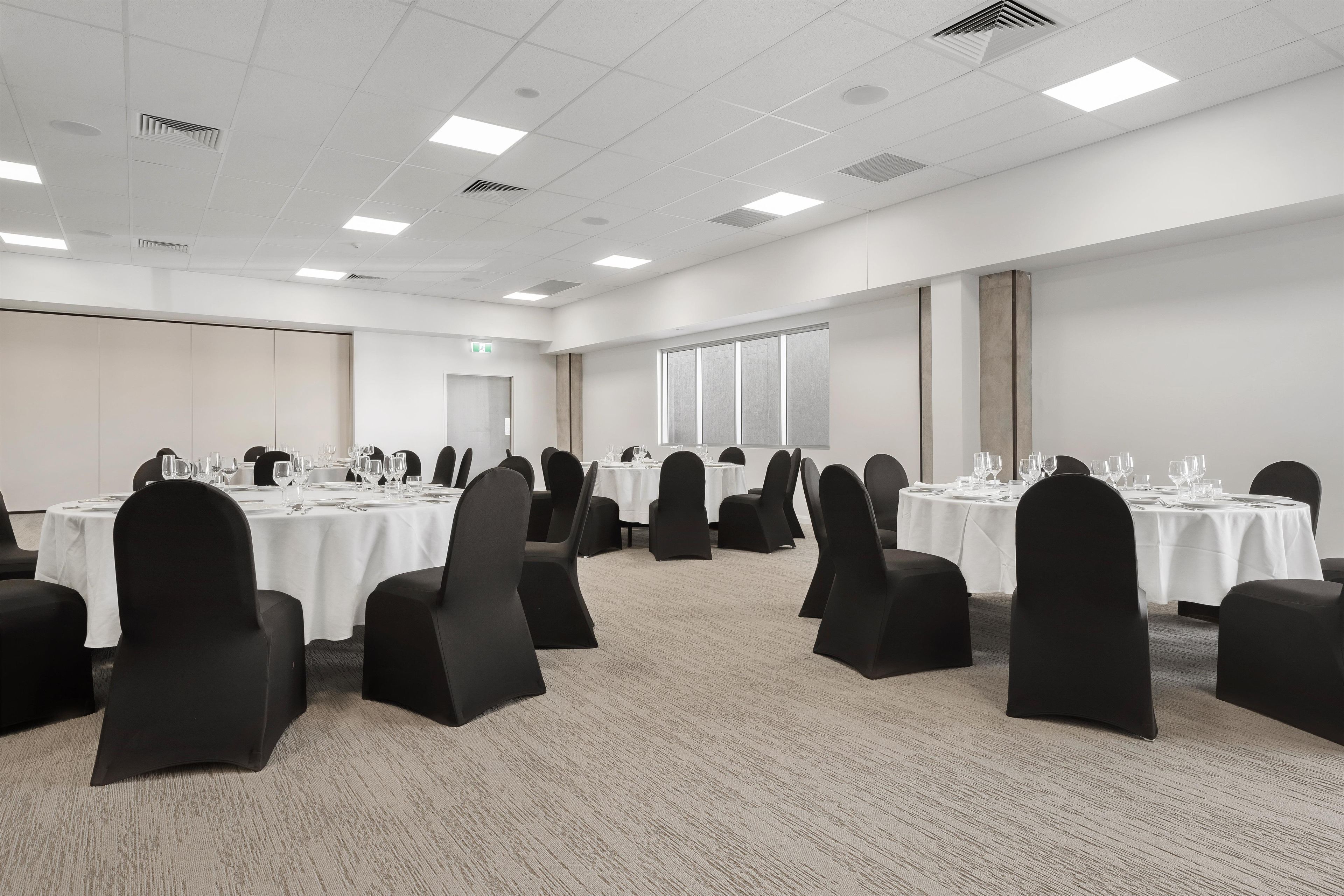A conference room with multiple tables, black chairs, and white tablecloths.
