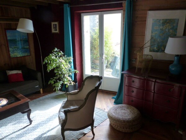 A cozy living room with a wooden floor, a dark-colored sofa, a chair, a lamp, a dresser, and a large window with curtains.
