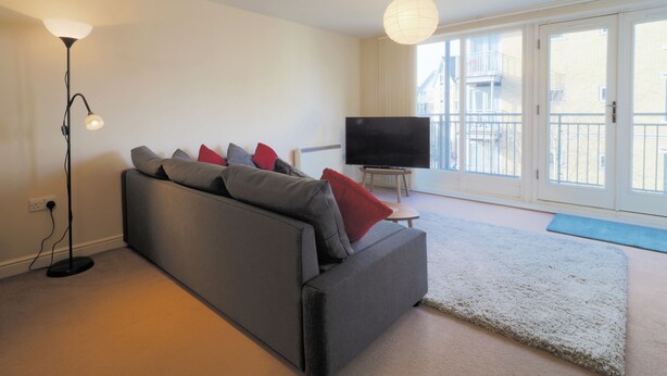 A modern living room with a grey sofa, red and grey cushions, a floor lamp, a TV on a stand, and a large window with a view of buildings. - Bingley Court (Canterbury)