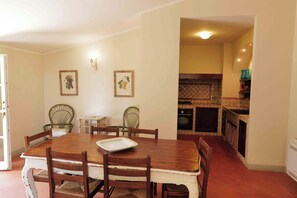 Uma sala de jantar com mesa e cadeiras de madeira, cozinha ao fundo e duas quadros na parede.