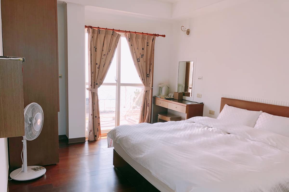 A hotel room with a bed, a desk, a chair, a fan, and a window with curtains.