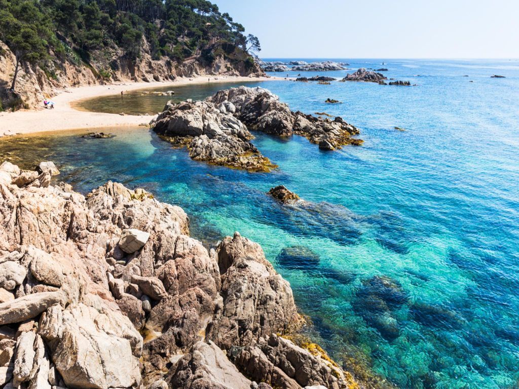 A coastal landscape with clear blue waters, rocky terrain, and a sandy beach.