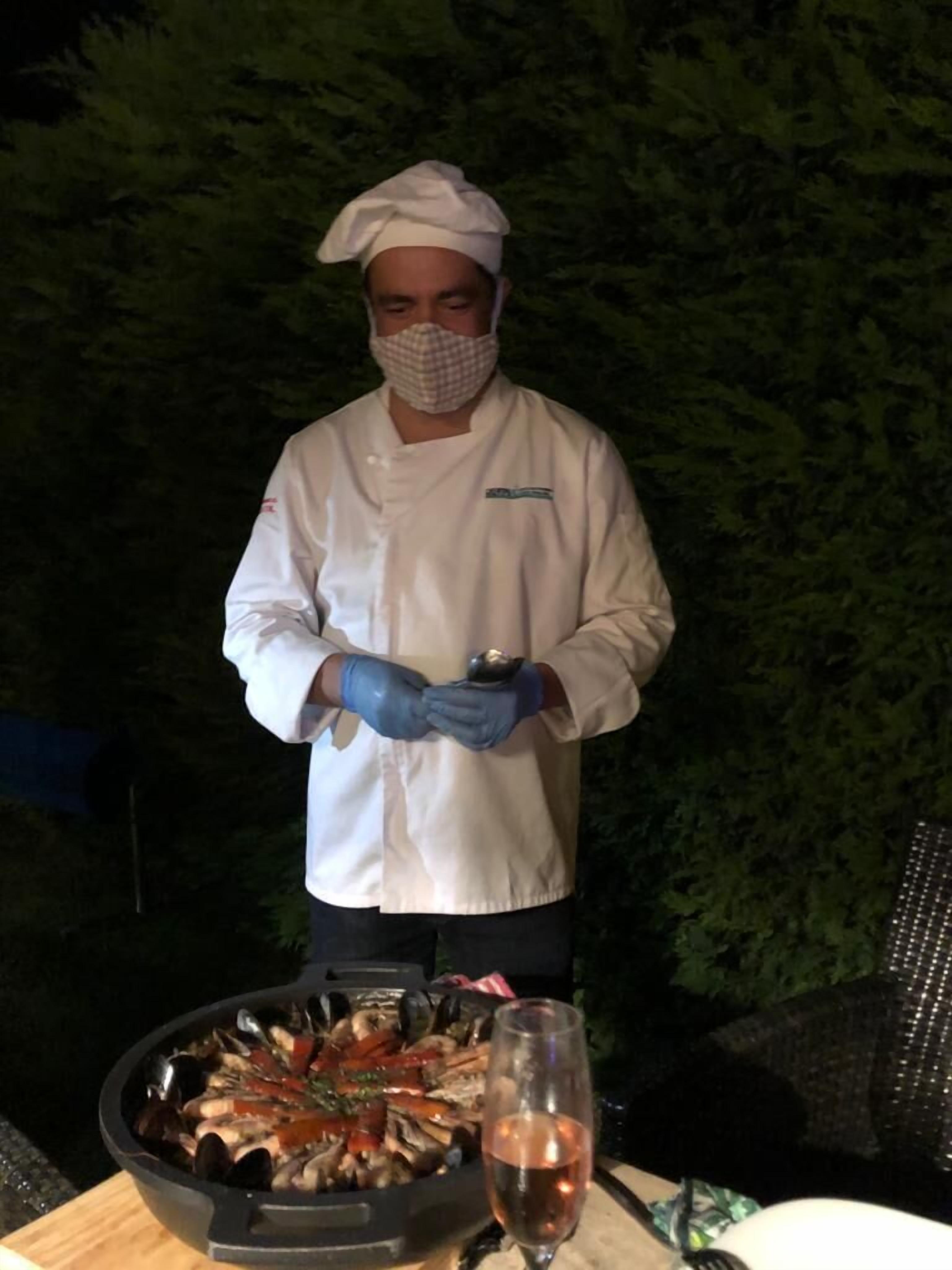 Een kok in een witte uniform met koksmuts en handschoenen, staand voor een grill met zeevruchten.