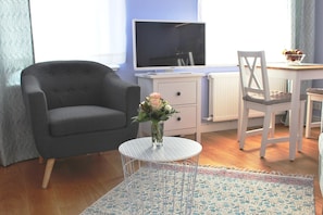 Una sala de estar con un sillón gris, una mesita redonda con un florero, un televisor blanco sobre un mueble bajo, un escritorio blanco con una silla y una alfombra estampada.