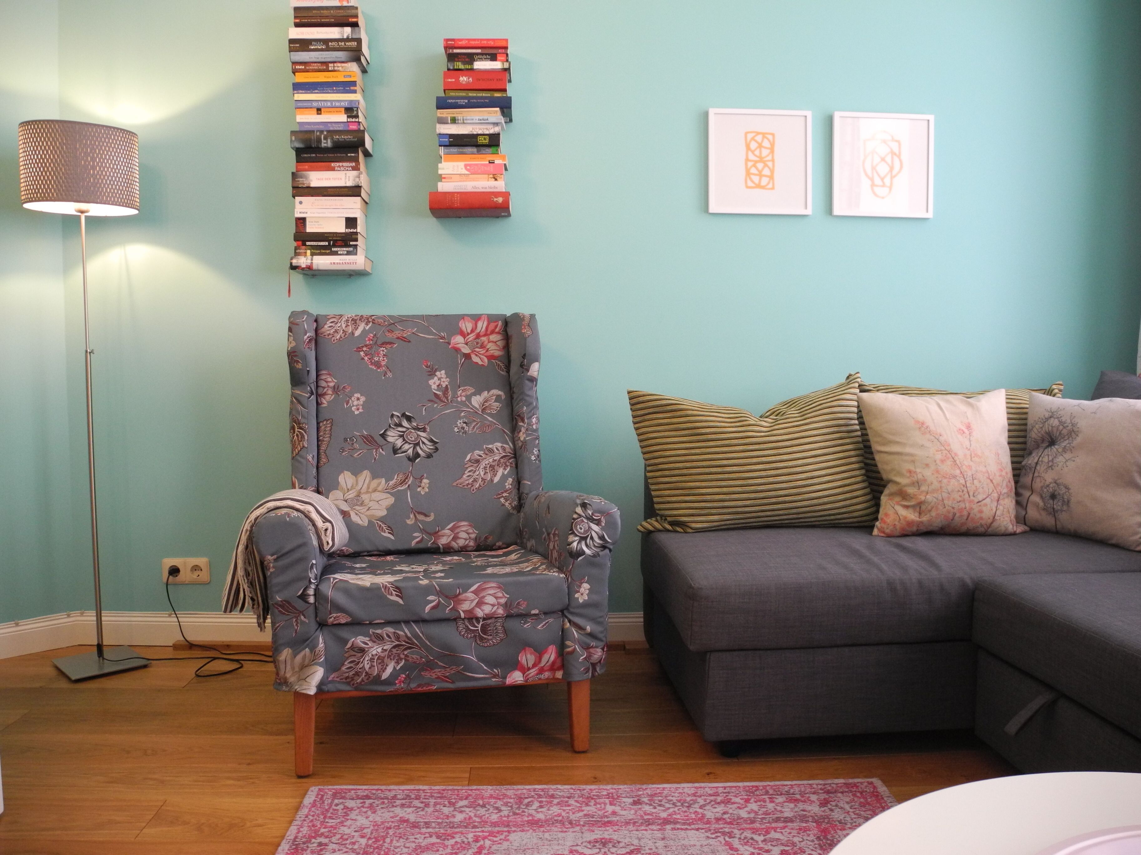 Ein Wohnzimmer mit einem blumigen Sessel, einem grauen Sofa und einem Bücherregal vor einer türkisblauen Wand.