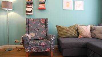 Ein Wohnzimmer mit einem blumigen Sessel, einem grauen Sofa und einem Bücherregal vor einer türkisblauen Wand.