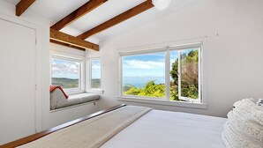 Une chambre dotée d’un grand lit, de deux fenêtres donnant sur un paysage verdoyant et des collines, et de poutres en bois au plafond.
