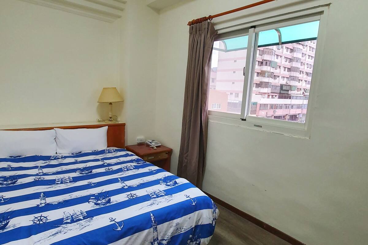A hotel room with a bed, a nightstand, a lamp, a window with curtains, and a view of buildings outside.