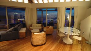 A modern living room with a sofa, two wicker chairs, a dining table, and large windows offering a city view.
