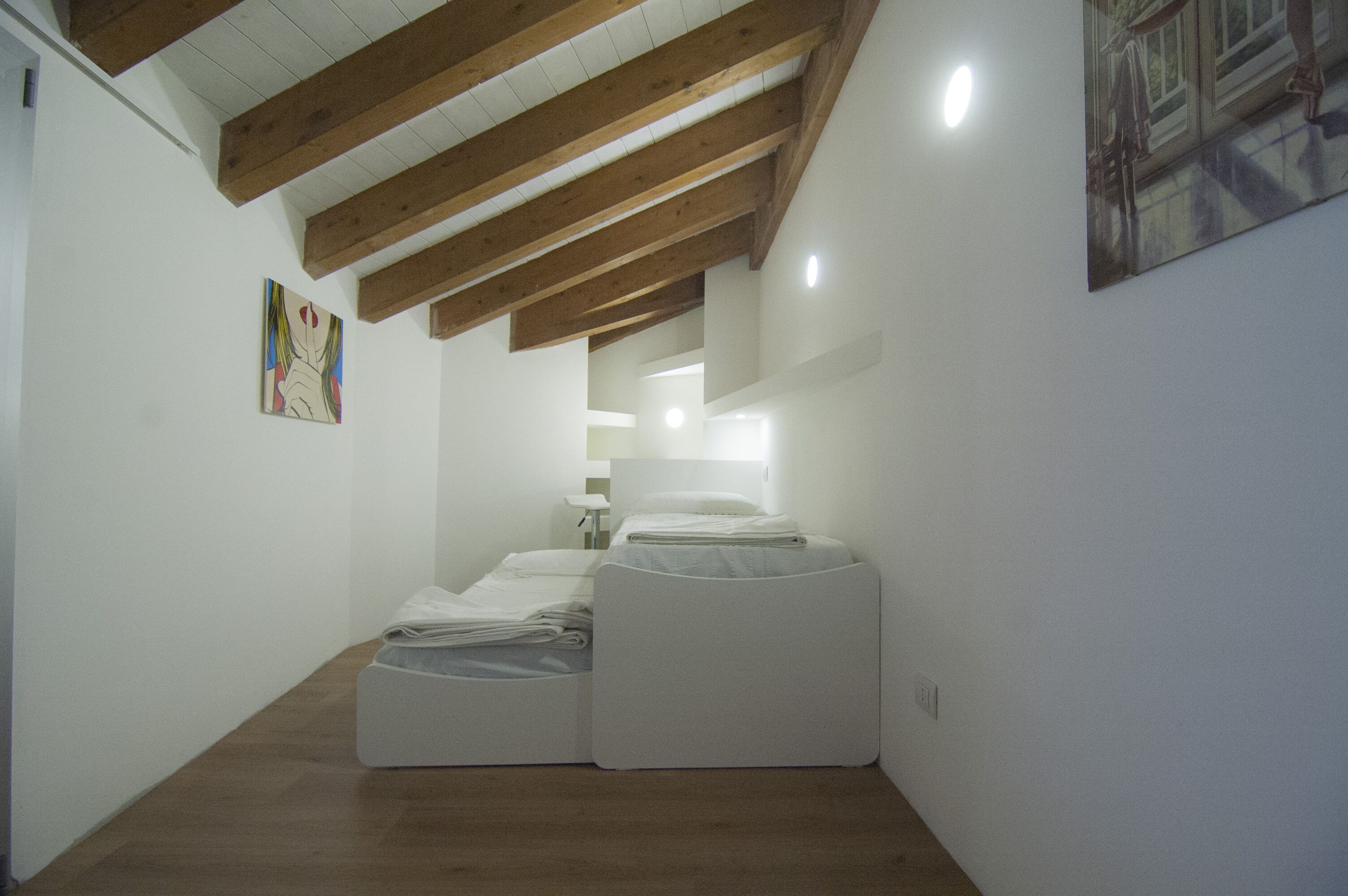 A narrow hallway with wooden beams on the ceiling, two white beds with folded linens, and a small table with a chair.