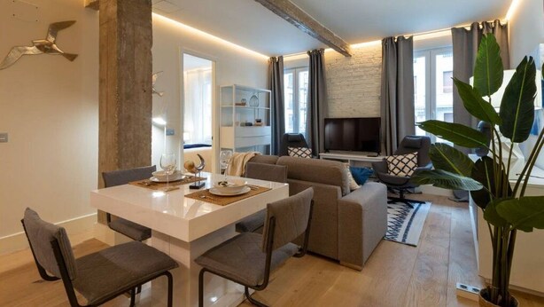 Ein modernes Wohnzimmer mit einem weißen Esstisch, grauen Sofas und einer großen Pflanze.